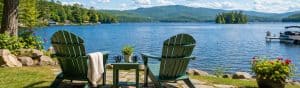 Two Adirondack chairs on a lakeside lawn with a view of calm water, trees, and mountains, perfect for recharging and enjoying nature's tranquility.