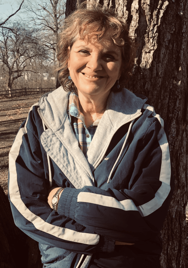 Lynn Sanders leans against a large tree as the sun shines down on her smiling face. She is wearing a light jacket on a crisp fall day