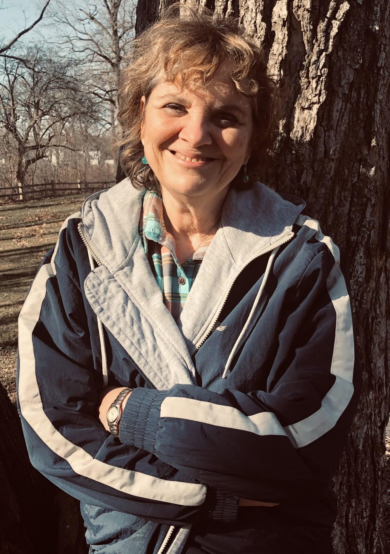 A woman smiling outdoors next to a large tree, dressed in casual and outdoor clothing, representing community engagement and nature appreciation.