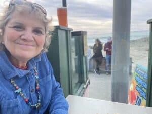 Bright smiling woman in a blue jacket at an outdoor beachside cafe with ocean view, wearing a colorful beaded necklace, on a cloudy day, promoting engaging community and coastal lifestyle with friendly atmosphere.