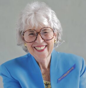 Stylish elderly woman with glasses smiling confidently, wearing a blue professional blazer and badge, symbolizing leadership, empowerment, and experience.