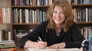 Energized woman author smiling at camera, seated at desk with books and journal, symbolizing storytelling expertise and leadership in media training, branding, and content creation.