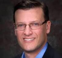 A professional headshot of a confident businessman with glasses, smiling, dressed in a suit, representing leadership, influence, and media excellence for branding.