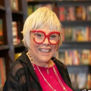 Energetic woman with blonde hair, red glasses, and a bright smile in a bookstore setting, promoting media solutions for impactful storytelling and brand growth.